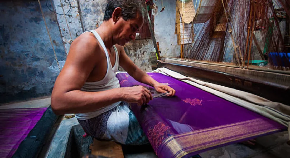 A woman draped in a vibrant silk saree stands against a backdrop of saree weaving looms, showcasing the intricate craftsmanship of traditional Indian sarees.